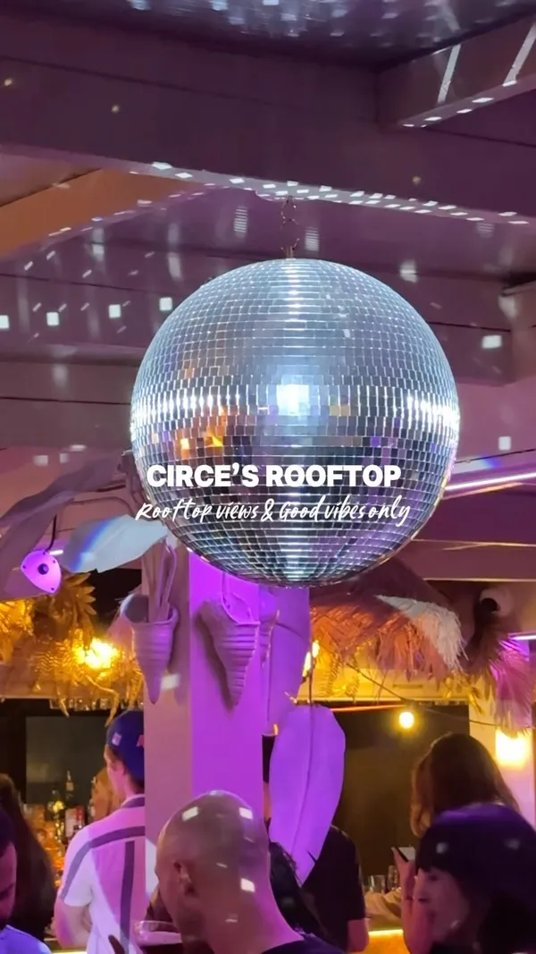 This video showcases Circe's Rooftop bar in Waterloo, London. The video features a disco ball, straw umbrellas, a woman talking on a phone, a bartender making drinks, various food dishes including fries, salad, and appetizers, and cocktails. The presentation emphasizes the bar's decor, including neon signs and themed lighting. The main focus is on the bar's atmosphere and offerings.