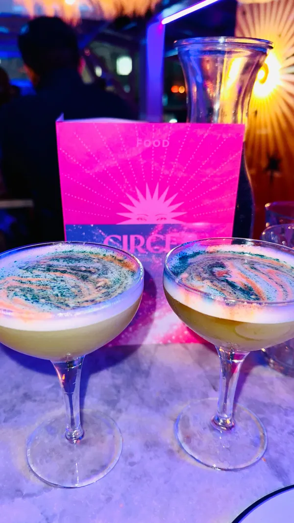 The video promotes Circe's Realm, a rooftop bar event in London. It begins with a shot of the entrance and a wall mural, followed by scenes of the bar's interior, featuring people socializing, a magician performing card tricks, and tarot card readings. The video showcases cocktails, sharing plates with grilled halloumi, salads, and mushroom dishes. It also shows a caricaturist drawing guests as mythical figures and a golden telephone prop. The main focus is on highlighting the unique experiences offered at the event, including cocktails, food, tarot readings, caricatures, and a chance to win prizes.