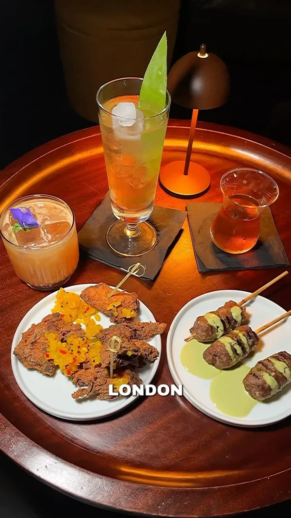 The video showcases a hidden cocktail bar located inside a hotel in London. It begins with a person approaching a door with a unique hand-shaped handle, then transitions to the bar's interior, which has a cozy, dimly lit ambiance with comfortable seating. The video features close-ups of various cocktails with unique garnishes and small food bites, including meat skewers and fried chicken with relish. Text overlays highlight the bar's concept of globally-inspired drinks and tasty bites. The video focuses on the bar's unique theme, drinks, food, and intimate atmosphere.