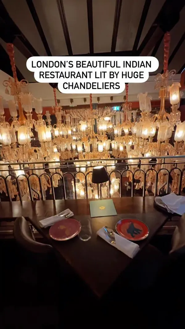The video showcases Colonel Saab, an Indian restaurant in Trafalgar Square, London. It features the restaurant's interior with large chandeliers and ornate decor. The video highlights a 5-course set menu inspired by 'Clueless the Musical', showing various dishes including a cocktail with a flower garnish, different curries with rice, a chicken dish, a pastry appetizer, and a cheesecake dessert. The food is presented on colourful plates. The video focuses on the ambiance and the set menu offering.