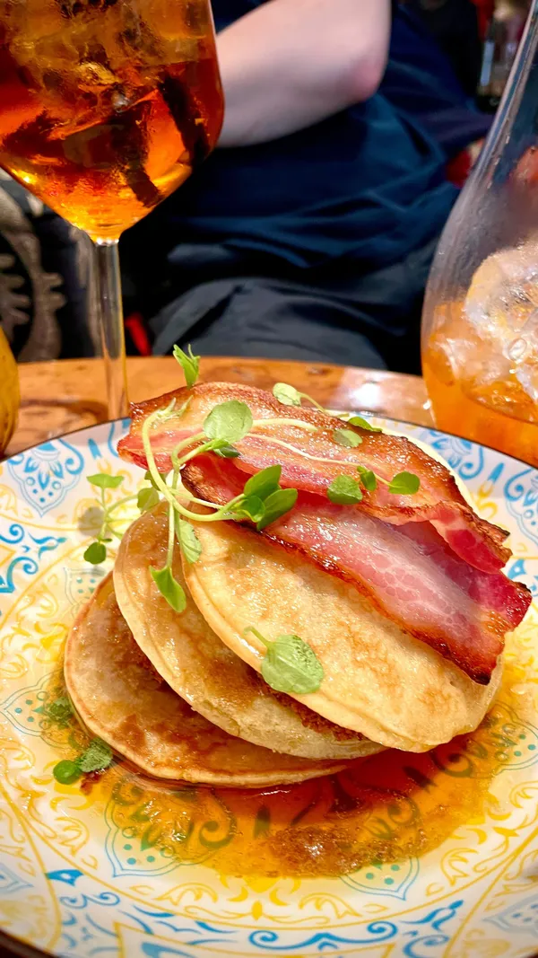 This video showcases a bottomless brunch at The Little Orange Door in Clapham, London.  The bar's interior is highlighted, resembling a house with various rooms. Food items featured include pancakes with bacon and a chicken burger with fries.  Aperol spritzes and other cocktails are shown. The video emphasizes the fun, social atmosphere of the brunch and the venue's unique design.