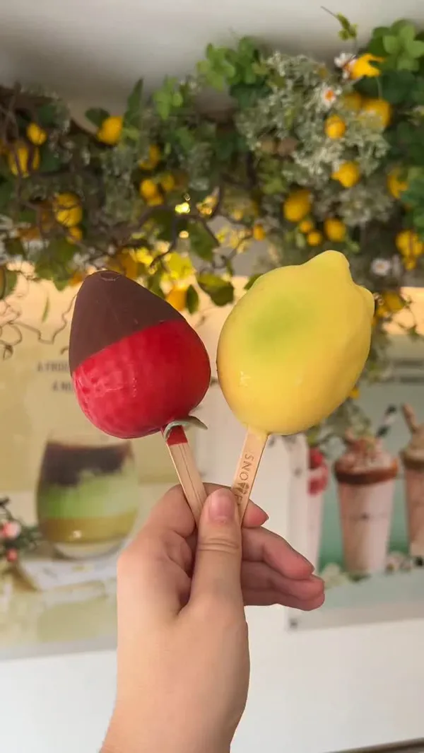 The video showcases Snowflake Gelato, a gelato shop in London. The main focus is on their unique fruit-shaped gelato popsicles, specifically the 'Snow Lemon' (lemon-shaped) and 'Snow Berry' (strawberry-shaped). The video shows these products being selected from a display case, with the 'Snow Berry' being dipped in melted chocolate. The creator then shows close-ups of the finished products and takes bites to reveal the gelato inside. The voiceover describes the flavors, mentions the use of quality British ingredients (Jersey milk and Somerset cream), and highlights other available desserts like sundaes, waffles, and different gelato flavors.