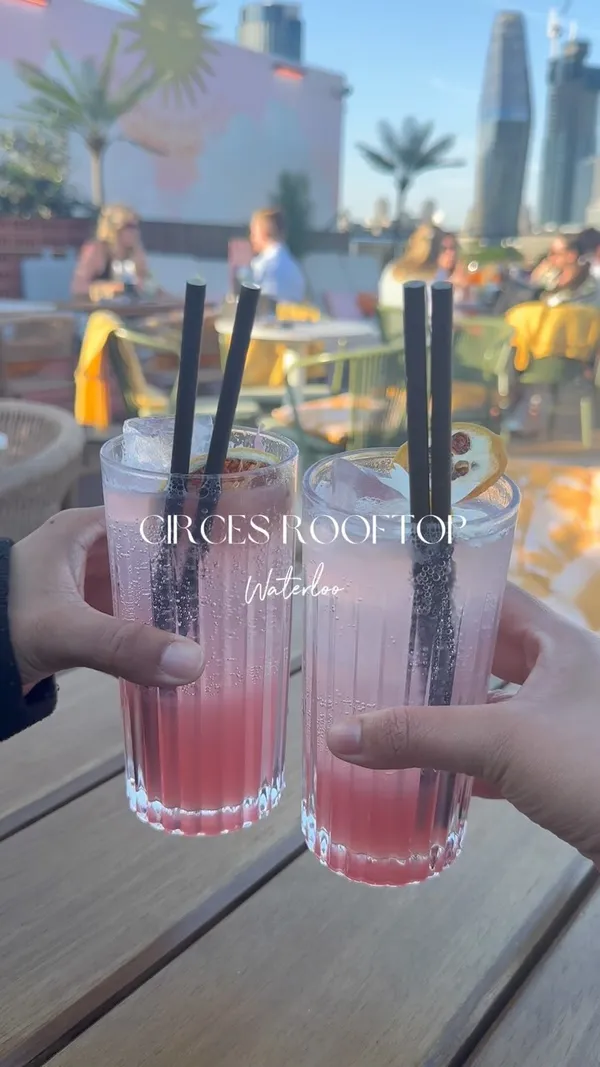 The video showcases Circes Rooftop in Waterloo, London. It features shots of the outdoor seating area, the drinks and food menus, two pink cocktails with garnishes, and a spread of food including hummus with pita bread, meat skewers, french fries, calamari, and a shrimp dish. The setting is an outdoor rooftop with views of the city skyline in the background.
