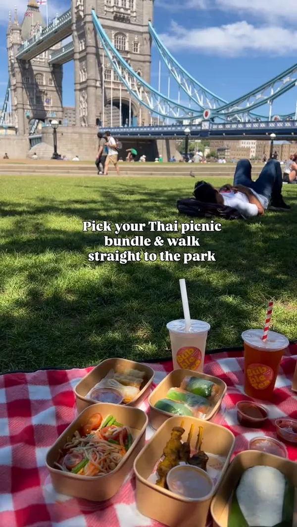 The video promotes a picnic bundle from Rosa's Thai restaurant. It begins with a view of the restaurant's exterior, then transitions to a scenic picnic spot in a park with Tower Bridge in the background. The video showcases the contents of the picnic bundle, which includes chicken satay, summer rolls, papaya salad, sticky rice, and bubble teas, all laid out on a red and white checkered blanket.