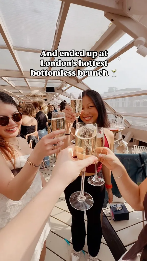 The video showcases a bottomless brunch experience at a rooftop venue. It begins with a shot of the venue entrance featuring a large mural. It then transitions to close-ups of multiple glasses of champagne, followed by people toasting with drinks. The video includes shots of performers in elaborate costumes, guests enjoying Mediterranean small plates (including salads and dips), and a lively atmosphere with people at the bar and tables. Text overlays provide context about the event being a bottomless brunch with cocktails and food.