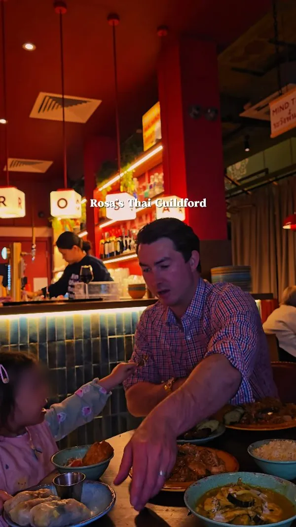 The video showcases a Thai restaurant experience at Rosa's Thai Guildford. It features various Thai dishes including fried rice, skewers, spring rolls, and fresh spring rolls with dipping sauce. It also shows a drink in a mason jar with a straw, and a toast with a beer glass. The video includes shots of the restaurant exterior, interior decor (neon sign, bar area), and the influencer and her child enjoying the meal. Text overlays identify the restaurant and mention a 

20 per person set menu.