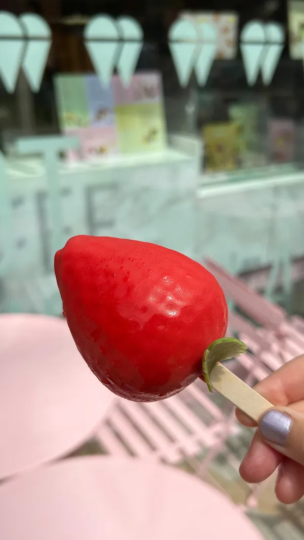 The video showcases gelato popsicles from Snowflake Gelato. It opens with a shot of chocolate sauce being stirred, followed by a display of three different gelato treats in a pink box: a strawberry-shaped popsicle, a lemon-shaped popsicle, and a rectangular chocolate-covered bar with pistachio drizzle. The video then shows close-ups of each treat, including shots of the chocolate bar and lemon popsicle being broken open to reveal their gelato fillings.