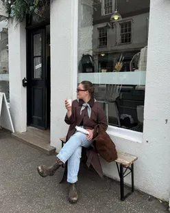 Person wearing a neckerchief and trench coat, enjoying the charming shops and scenery of Dunkeld, Scotland.