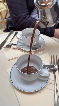 Enjoying iconic hot chocolate at Carette in Paris with a friend, featuring whipped cream and a cozy café atmosphere.