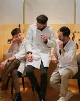 A polished, well-lit lifestyle photograph featuring three individuals in lab coats participating in a perfume-making workshop. The subjects are engaged in the activity, smelling scent strips, which effectively communicates the nature of the experience at the Experimental Perfume Club. The composition is clean and professional, with a neutral, minimalist background that keeps the focus on the participants and the activity.