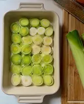 A clean, overhead food photography shot featuring sliced leeks arranged neatly in a baking dish, likely in preparation for a roasted vegetable recipe. The lighting is soft and natural, highlighting the texture and vibrant green tones of the produce, while the composition is balanced and professional.
