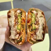 A high-quality, close-up shot of a chicken sandwich held in hand, effectively showcasing the textures of the fluffy focaccia, blackened chicken, avocado, and sauces. The natural lighting and sharp focus make the food look fresh and highly appetising.
