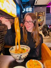 A portrait-style shot of a woman holding up a long, wide noodle with chopsticks from a bowl at a restaurant, with a plate of corn ribs visible in the foreground.