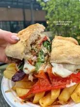 A close-up, handheld shot of a loaded steak sandwich served on a paper plate with fries, likely at a market stall. The focus is on the melted cheese and filling, highlighting the 'proper sarnie' appeal.