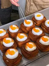 A high-quality, behind-the-scenes shot of a pastry chef finishing a batch of cube-shaped pastries topped with meringue and mandarin segments. The image captures the precision of the plating process in a clean, well-lit kitchen environment.