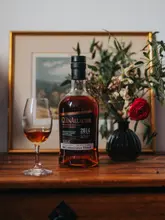 A high-quality, atmospheric product photograph featuring a bottle of The Glenallachie Sinteis Series single malt whisky alongside a glass of the spirit, set against a warm, home-style background with a framed picture and a vase of flowers.