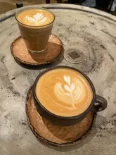 A high-angle, close-up shot of two coffees served in ceramic cups on a textured grey table. The latte art is clearly visible, and the lighting is soft and natural, highlighting the texture of the cups and the surface.