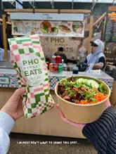 A first-person perspective shot taken at a market stall, featuring a bowl of Vietnamese food and a branded paper bag. The image captures the casual, authentic vibe of the street food vendor, though the framing is tight.