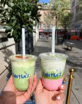 A bright, outdoor shot featuring two layered matcha lattes held up against a street background, showcasing the vibrant green and pink colours of the drinks.