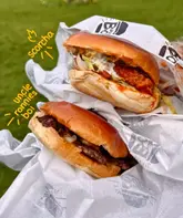 A casual, handheld shot of two burgers in branded takeaway paper wrappers, set against a blurred outdoor background. The image is a simple, functional snapshot intended to showcase the product for a review.