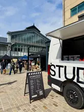 A documentary-style, eye-level shot of a coffee van at an outdoor market, featuring a menu board in the foreground and market stalls in the background.