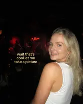 A candid-style portrait of a woman posing in a dimly lit jazz club or bar, with a live band visible in the background. The image uses a harsh flash aesthetic typical of nightlife photography to capture a social moment.