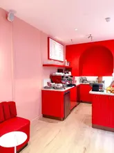 A wide-angle interior shot of a cafe in Mayfair, featuring a bold, monochromatic red and pink color scheme. The image captures the service counter and seating area, showcasing the venue's distinct aesthetic.