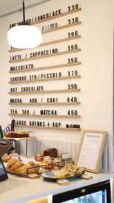 A short, montage-style video showcasing the interior and menu offerings of The Missing Bean cafe in Charlbury. The video features shots of the exterior, the menu board, the seating area, the bakery shelf, and close-ups of coffee, tea, a croissant, and a sandwich.