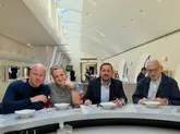 A high-quality, well-lit photograph of four individuals seated at a marble-topped table in a bright, modern restaurant setting. The image captures a professional or celebratory lunch atmosphere, with the subjects posed clearly for the camera.