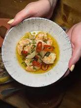 A close-up, first-person perspective shot of a bowl of garlic prawns being held by two hands, likely taken in a dimly lit restaurant setting using a direct camera flash.