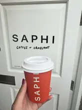 A simple, clean POV shot of a branded takeaway coffee cup held in front of the entrance to Saphi, a coffee and croissant shop in South London.