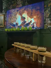 A close-up shot of a wooden flight board containing six shot glasses, positioned on a table in a dimly lit bar setting. The background features a fireplace screen and floral wallpaper, creating a cozy, moody atmosphere.