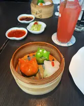 A close-up, high-angle shot of a bamboo steamer containing three colorful dim sum pieces, with a drink and side dishes visible in the background at Yauatcha City.