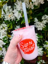 A close-up, eye-level shot of a hand holding a pink fizzy drink in a branded plastic cup, set against a backdrop of white flowers.
