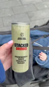 A short, handheld vlog-style video showing a visit to 'The Alcohol Company' off-license to purchase 'Stacked Drinks' matcha cans. The creator walks into the store, highlights the fridge display, and shows the product.