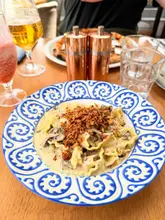 A top-down shot of a truffle mushroom mafaldine pasta dish served on a decorative blue and white plate, with a busy table setting in the background including a pizza, drinks, and salt and pepper shakers.