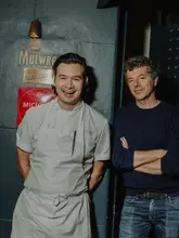 A portrait of a chef and another individual standing in front of a wall featuring a Michelin plaque, likely taken at Evelyn's Table. The image captures a candid, happy moment between the subjects.
