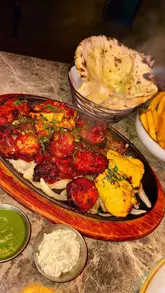 A food-focused reel showcasing a large mixed grill sizzler platter, cheesy garlic naan, masala fries, and drinks at De Baga in Chapel Allerton. The video uses close-up shots to highlight the food's texture and presentation, including a cheese pull and the sizzling effect of the platter.