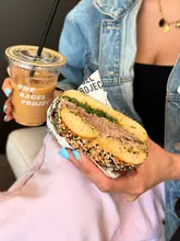 A casual, first-person perspective shot of a person holding a bagel and an iced coffee, likely taken inside a cafe. The image focuses on the product being consumed, typical of a social media review or lifestyle post.
