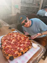 A casual, outdoor shot featuring a person posing with a very large pepperoni pizza, accompanied by a beer and a side of garlic dip. The setting appears to be a relaxed, semi-outdoor venue.
