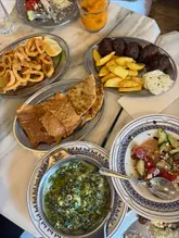 A top-down, overhead shot of a Greek mezze feast spread across a marble table, featuring plates of calamari, keftedes with fries, pita bread, spinach dip, and a Greek salad.