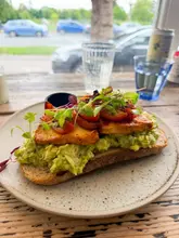 A close-up, eye-level shot of an avocado toast dish topped with grilled halloumi and roasted tomatoes, served on a rustic plate at a cafe.