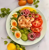 A high-quality, overhead food photograph of a prawn and egg salad, styled with fresh herbs and ingredients scattered around the plate on a clean, white marble surface.