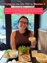 A photo of a creator smiling at a table in a Vietnamese restaurant, holding a cocktail, with various dishes spread out in front of her.