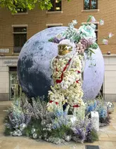 A high-quality, well-lit photograph of a floral astronaut sculpture surrounded by flowers and disco balls, part of the Chelsea in Bloom festival. The image captures the intricate detail of the floral arrangement against a backdrop of a moon prop and a brick building.