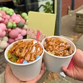 A bright, high-quality close-up shot of two cups of frozen yogurt held in hands, showcasing generous toppings and sauces. The background features a branded sign and a balloon arch, providing a clear, inviting context for the venue.