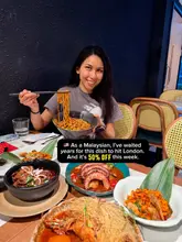 A promotional image featuring a creator holding a bowl of noodles at a restaurant table filled with various Malaysian dishes.