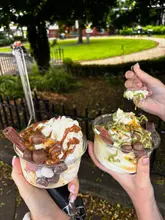 A close-up, eye-level shot of two hands holding loaded cups of frozen yogurt, topped with various chocolates, sauces, and fruit, set against a blurred park background.