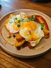 A top-down shot of a brunch dish featuring two fried eggs served over a bed of crispy potatoes with a side of spicy salsa brava sauce, garnished with pea shoots.