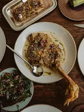 A high-quality, overhead shot of a 'malai murgh' chicken leg dish served at a restaurant. The dish is plated on a patterned plate with a creamy sauce and garnished with edible flowers. The lighting is moody and atmospheric, highlighting the textures of the food.